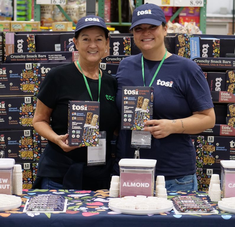 The mom-daughter co-founders of Tosi standing behind a demo with Tosi products and promotional materials.